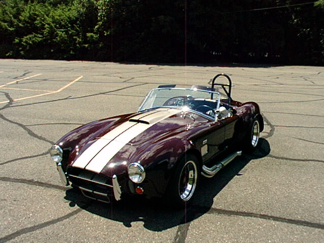 Maroon Cobra roadster with silver stripes, chrome wire wheels and side pipes in a parking lot
