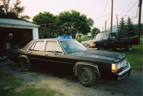 1989 Ford Crown Victoria LTD - Two vehicles, a black sedan and a black pickup truck, are parked outside a garage. The sedan appears to be a 1989 Ford Crown Victoria LTD.