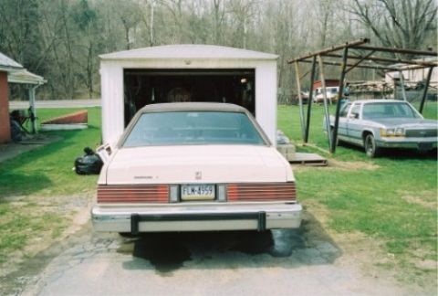 1987 Mercury Grand Marquis - A light-colored sedan is parked in front of a garage, with another vehicle visible to the right.