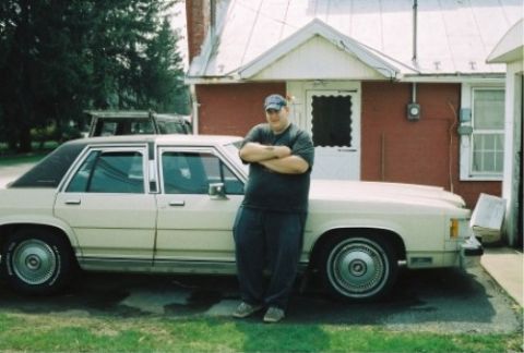 1987 Mercury Grand Marquis - A man poses next to a cream-colored sedan, with another vehicle in the background.
