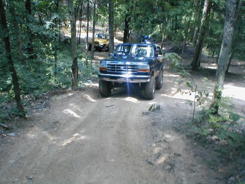 1995 Ford Bronco - A dark blue SUV drives on a wooded dirt path, with another vehicle in the distance.