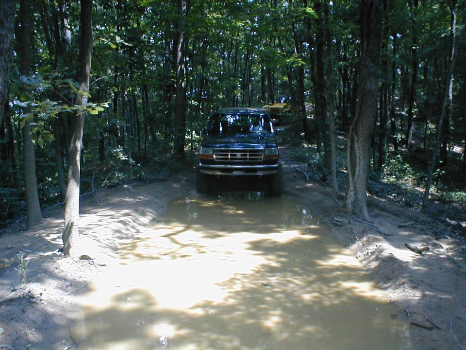 1995 Ford Bronco driving through a muddy puddle on a dirt trail in the woods