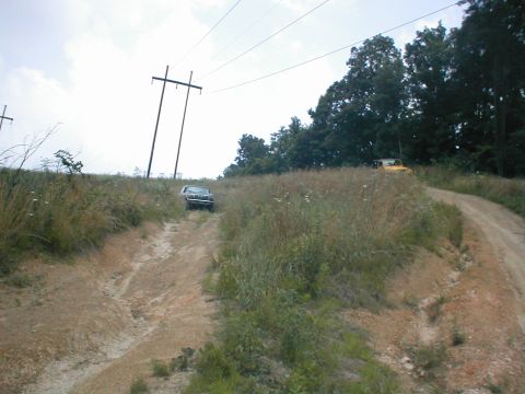 1995 Ford Bronco - A dark SUV navigates a dirt road with a yellow vehicle visible further up the path.