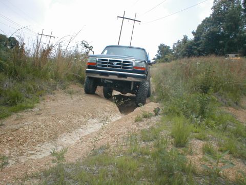 1995 Ford Bronco - 1995 Ford Bronco navigates an off-road trail with a deep rut ahead.