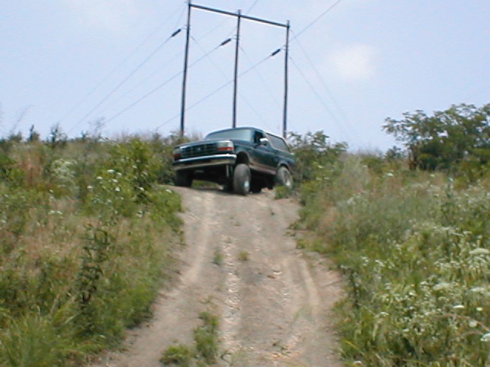 Dark green 1992 Ford Bronco driving up a dirt hill, front 3/4 view. Off-road tires, some vegetation on the sides