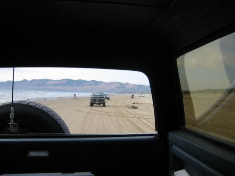 1985 Ford Bronco - A black 1985 Ford Bronco drives along a sandy beach, with mountains in the background and other vehicles present.