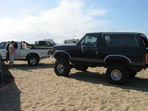 1985 Ford Bronco - A lifted black Ford Bronco is parked on a sandy beach next to a white pickup truck.