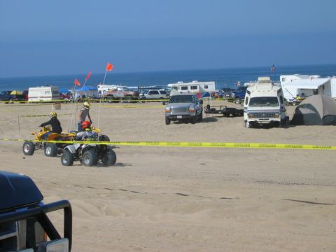1985 Ford Bronco - People ride ATVs on a sandy beach with RVs and campers parked nearby under a clear blue sky.