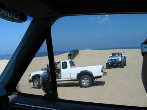 1985 Ford Bronco - Multiple trucks and an off-road vehicle are seen on sand dunes near the ocean.