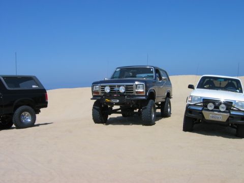 1985 Ford Bronco - Three modified off-road vehicles are parked on sand dunes with a clear blue sky above.