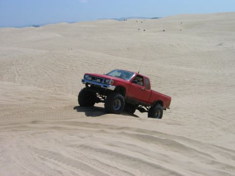 1985 Ford Bronco - A red pickup truck navigates a sandy terrain with a clear blue sky overhead.