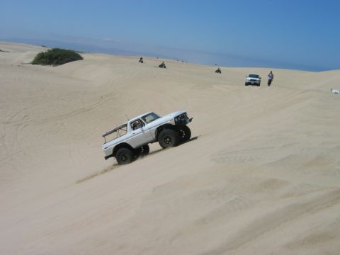 1985 Ford Bronco - A white Ford Bronco navigates a sandy incline, with other vehicles and people visible in the background.