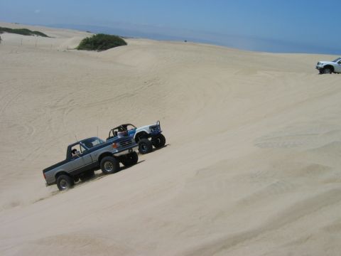 1985 Ford Bronco - Two off-road trucks navigate steep sand dunes under a clear blue sky.
