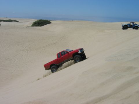 1985 Ford Bronco - Two off-road vehicles navigate sandy terrain under a clear blue sky.