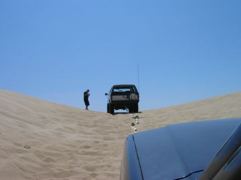 1985 Ford Bronco - A dark SUV is parked on a sand dune with a person standing next to it, while another vehicle is in the foreground.