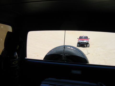 1985 Ford Bronco - A red pickup truck navigates sandy terrain, viewed through the rear window of another vehicle.