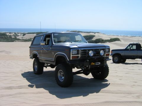 1985 Ford Bronco - A lifted Ford Bronco sits on sand dunes near the ocean, with another truck in the background.