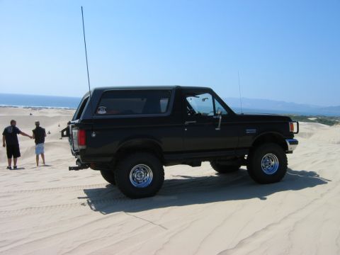 1985 Ford Bronco - A black 1985 Ford Bronco is parked on sand dunes near the ocean, with two people standing in the background.