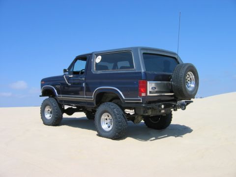 1985 Ford Bronco - This lifted Ford Bronco is parked on a sandy terrain under a clear blue sky.