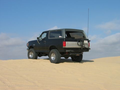 1985 Ford Bronco - This 1985 Ford Bronco is shown from the rear, parked on a sandy terrain under a blue sky.