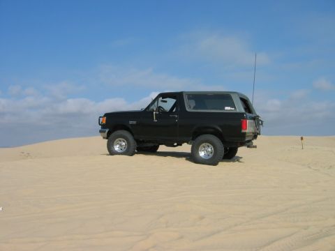 1985 Ford Bronco - A black Ford Bronco is parked on a sandy terrain with a clear blue sky above.