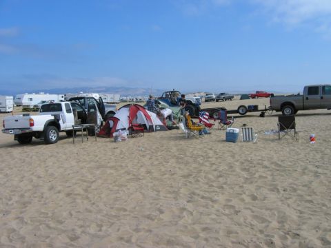 1985 Ford Bronco - A beach campsite is set up with a white pickup truck and tents, with other vehicles and RVs visible in the distance.