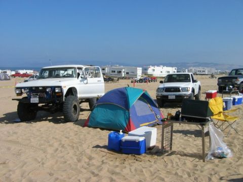 1988 Ford Bronco - Beach campsite with a heavily modified 1970s Ford truck, a tent, and other vehicles under a clear blue sky.