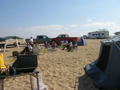 1988 Ford Bronco - A group of people are gathered around a campfire on a sandy beach, with several trucks and campers in the background.