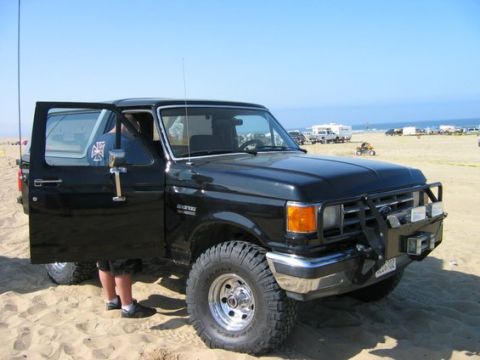 1988 Ford Bronco - A black 1988 Ford Bronco parked on the sand with its driver's door ajar, showing off its off-road tires and front brush guard.