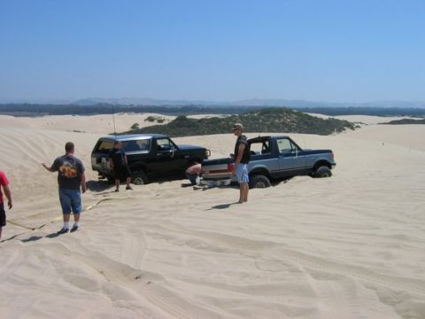 1988 Ford Bronco - Two trucks are stuck in the sand dunes, with people attempting to free them.