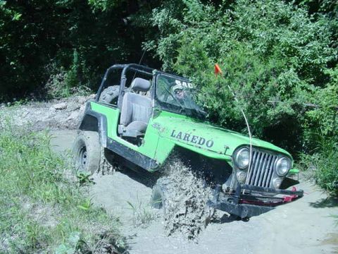 1992 Ford Bronco - Off-roading Jeep Wrangler navigating a muddy trail surrounded by dense foliage.