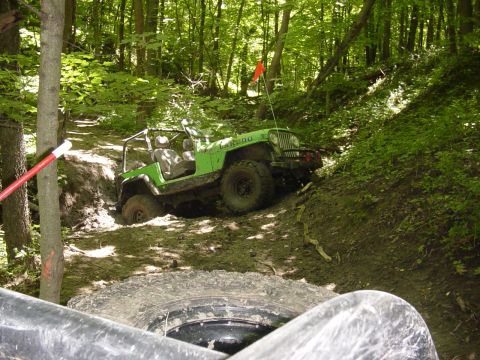 1992 Ford Bronco - A green Jeep navigates a challenging muddy incline on a wooded trail, with another vehicle's tire visible in the foreground.
