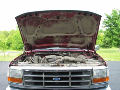 1995 Ford Bronco - The front grille of a red 1995 Ford Bronco is visible with the hood open, revealing a dusty engine compartment.
