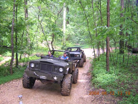 1984 Chevrolet Blazer - Two olive green off-road vehicles drive down a dirt path in a wooded area.