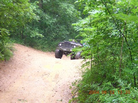 1984 Chevrolet Blazer - Olive green military-style jeep with large tires driving on a dirt road through a forest.