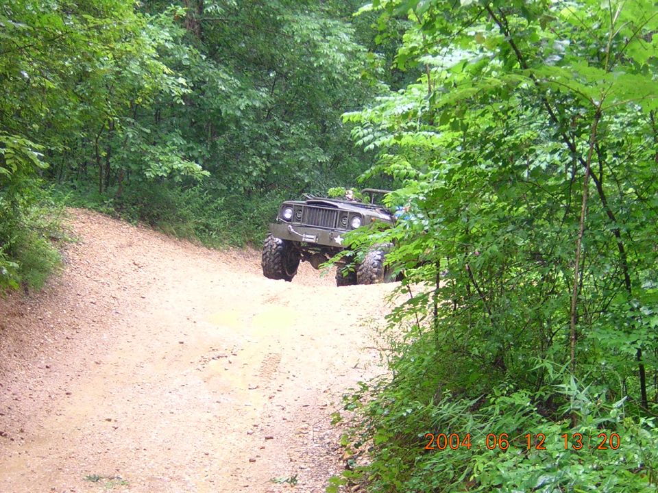 Olive green military-style jeep with large tires driving on a dirt road through a forest
