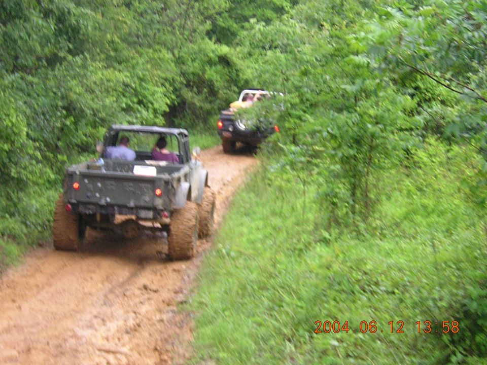Two off-road vehicles driving on a muddy dirt path through a wooded area