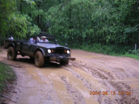 1984 Chevrolet Blazer - Jeep Gladiator J-series pickup truck navigates a muddy off-road trail with trees in the background.