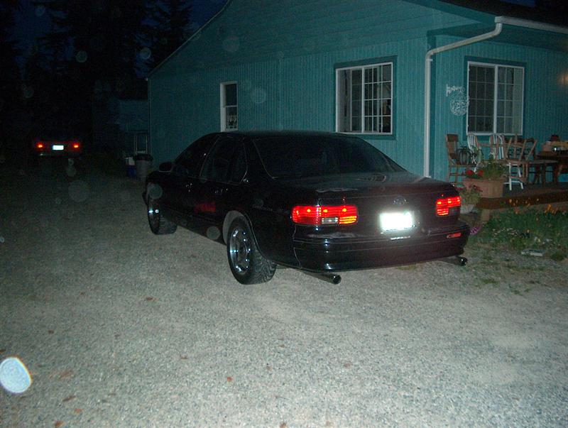 Black sedan parked on gravel at dusk, rear view with tail lights lit near teal house