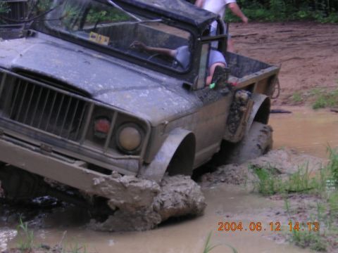 1984 Chevrolet Blazer - A muddy Jeep CJ-5 navigates a treacherous off-road path, its front-left wheel submerged in thick mud.