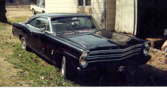 Black 1960s Ford fastback coupe parked beside a shed with a pickup in the background