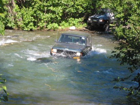 1985 Ford Bronco - Two SUVs navigate a shallow river surrounded by lush green foliage.
