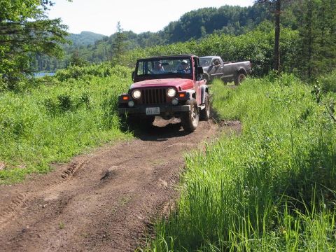 1985 Ford Bronco - Two vehicles, a Jeep Wrangler and a pickup truck, navigate a dirt path in a wooded area.