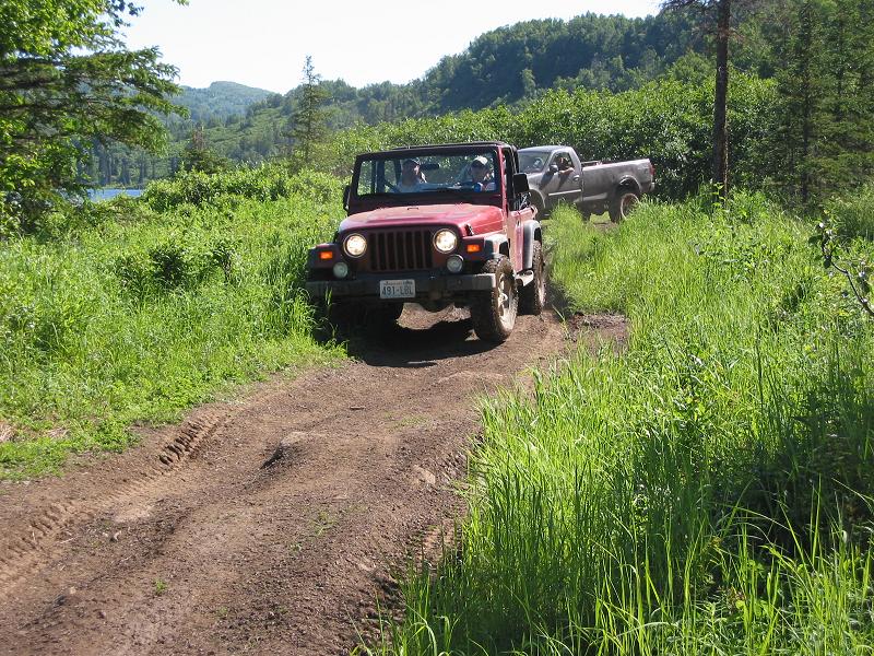Topless red Jeep Wrangler leading a gray pickup down a grassy dirt trail near a lake