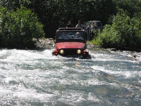 1985 Ford Bronco - A group of people are off-roading in a red Jeep Wrangler as it navigates through a shallow river.