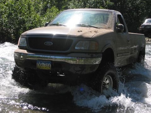 1985 Ford Bronco - A tan Ford F-150 navigates a water crossing, with another vehicle visible in the background.