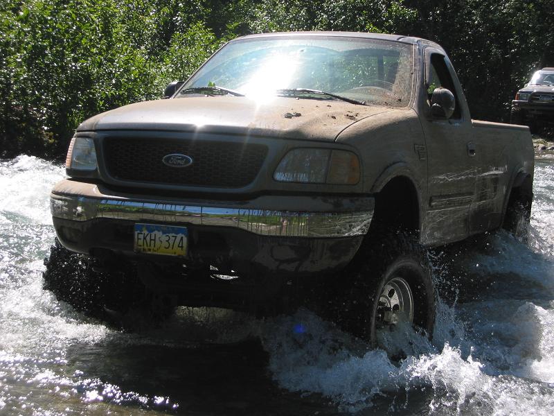 Muddy tan Ford regular cab pickup driving through a stream, water spraying at the bumper