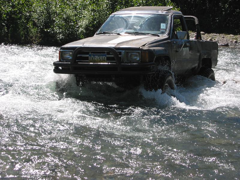 Mud-covered lifted pickup with brush guard and snorkel crossing a river through whitewater
