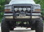 1993 Ford Bronco - Very small, low-resolution front view of a lifted 1990s Ford truck or SUV with a row of round auxiliary driving lights across a light bar a…