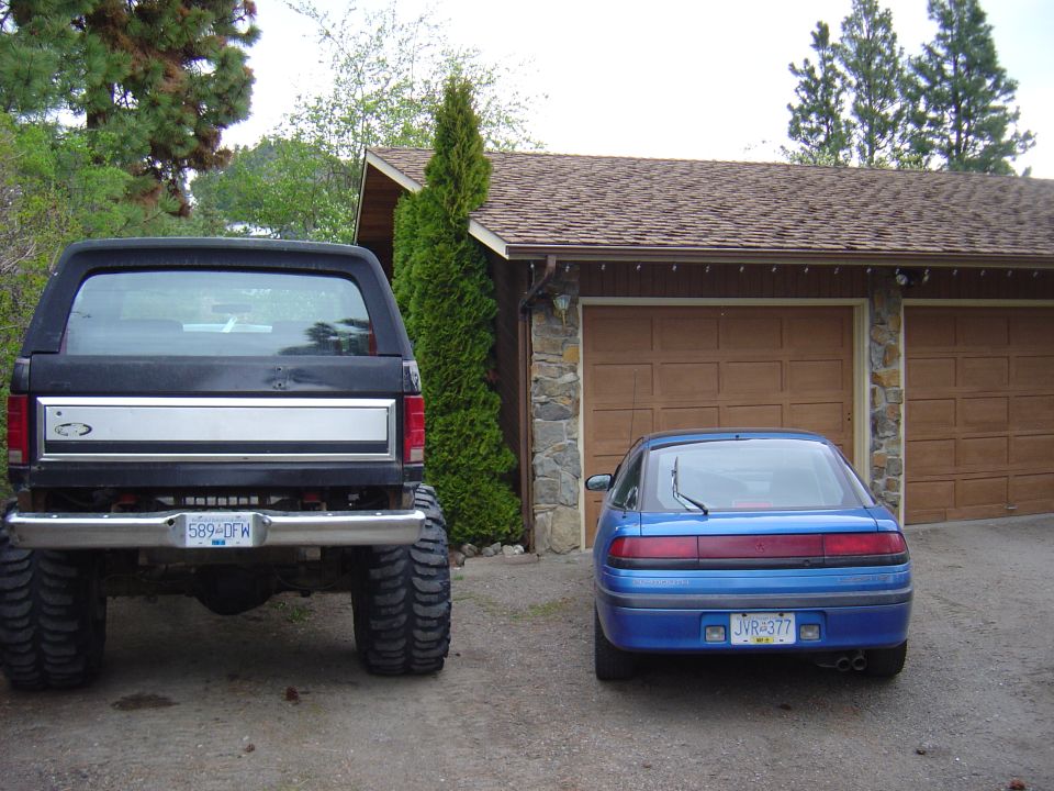Lifted black Ford Bronco with huge tires parked next to a blue Mitsubishi hatchback by a garage